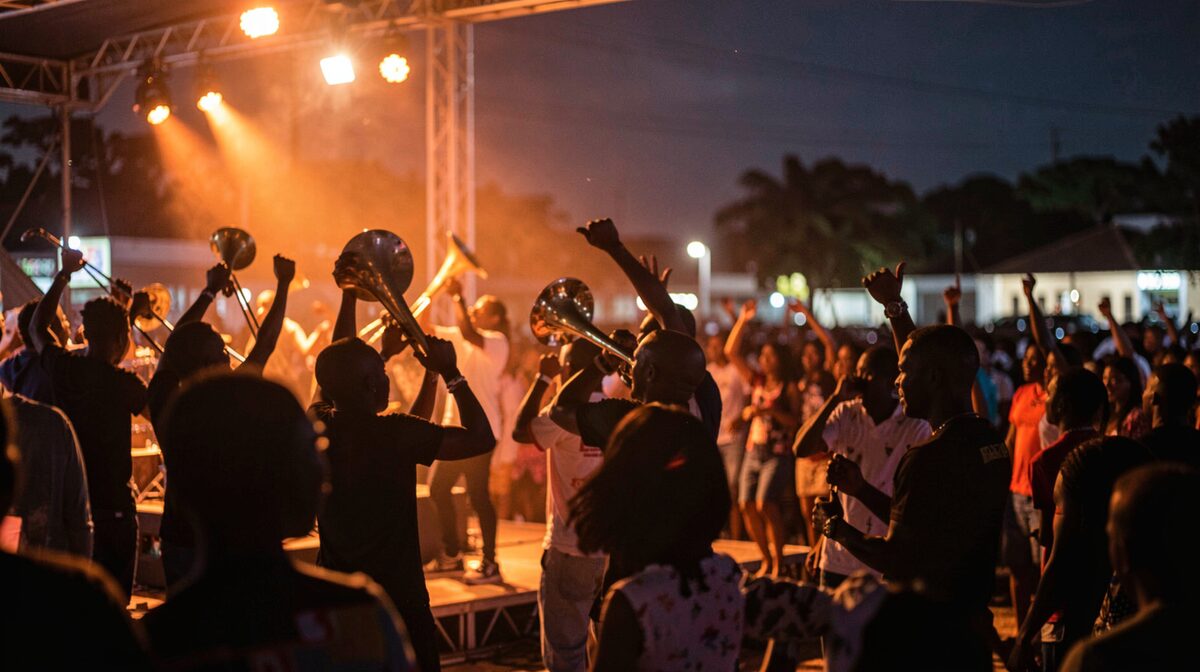 Concert en plein air la nuit, foule compacte bras levés face à une scène éclairée de projecteurs orangés, silhouettes d’une section de cuivres au premier plan